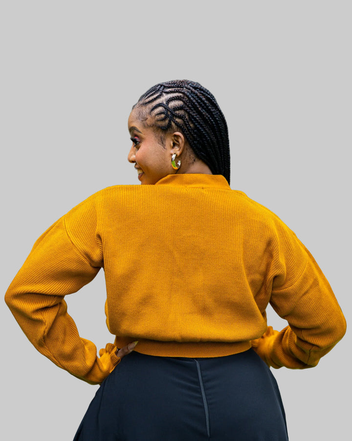 Woman wearing a mustard yellow sweater with braided hair, standing outdoors with greenery in the background.